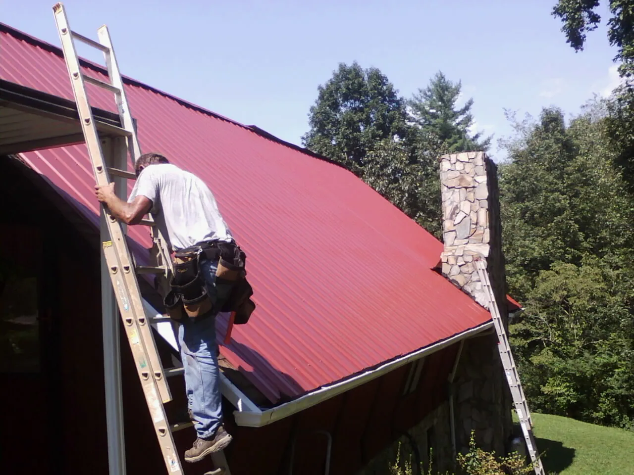 Roofer on a ladder beside a red metal roof and stone chimney — Soffit & Fascia Repair by Roofing Brothers in Round Hill
